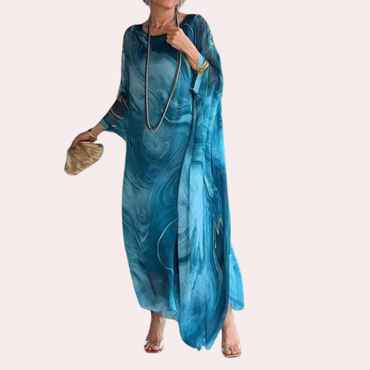 Vrouw in elegante blauwe kaftan jurk met abstract patroon, lange ketting en gouden armband. Perfect voor zomer, strand of casual chique evenementen.