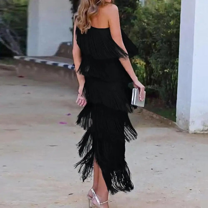 Vrouw in elegante zwarte franjesjurk met asymmetrische schouder, draagt zilveren clutch en hoge hakken, buiten bij een tuinpad.