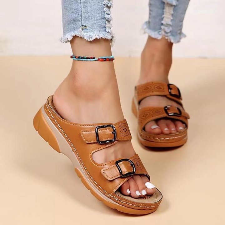 Bruine leren sandalen met dubbele gespen, gedragen door een persoon met een enkelbandje en gescheurde jeans. Comfortabele zomerschoenen voor dames.