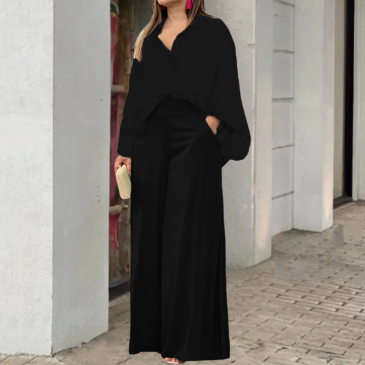 Vrouw in zwarte elegante jumpsuit met wijde pijpen, roze oorbellen en beige clutch, staand op een stedelijke achtergrond. Perfect voor avondmode.