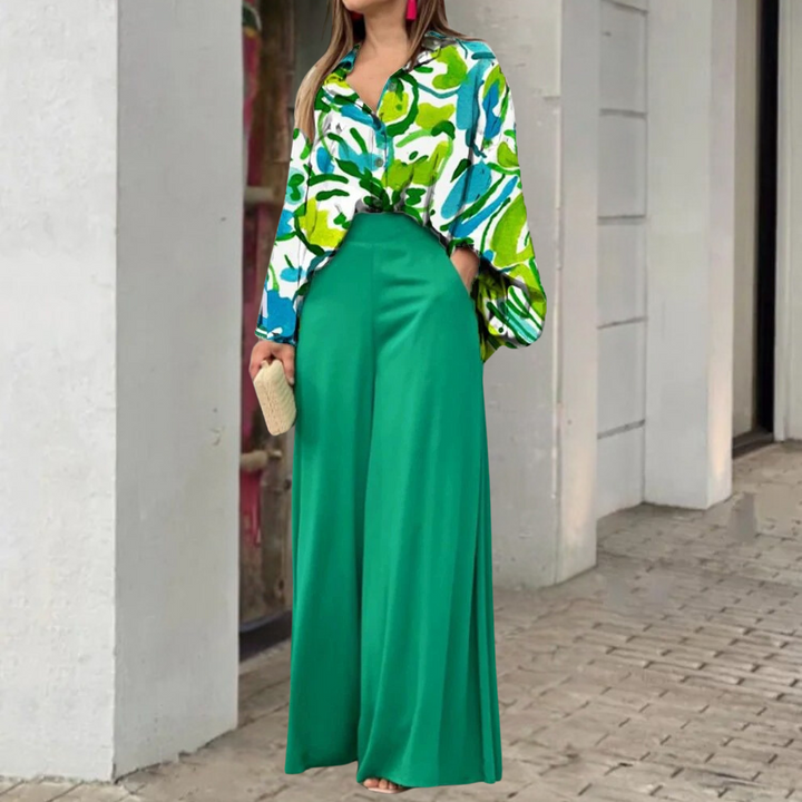 Vrouw in groene wijde broek en kleurrijke blouse met abstract patroon, staand op stoep. Modieuze outfit, trendy dameskleding, stedelijke achtergrond.
