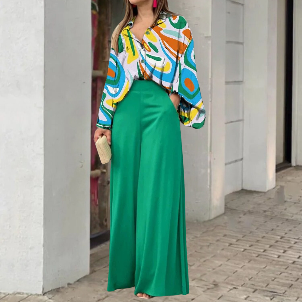 Vrouw in kleurrijke blouse met abstract patroon en groene wijde broek, staand op stoep. Modieuze outfit, trendy dameskleding, stedelijke achtergrond.
