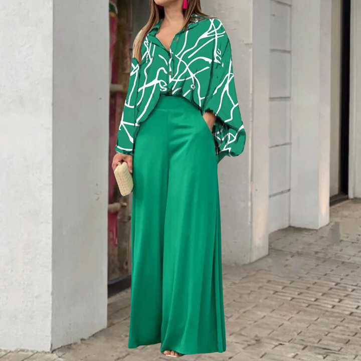 Vrouw in groene blouse met abstract patroon en wijde groene broek, houdt een beige clutch vast. Stijlvolle mode, elegante dameskleding.