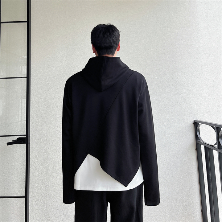 Man in zwarte asymmetrische hoodie met capuchon, witte onderlaag zichtbaar, moderne minimalistische stijl, staand voor witte muur.