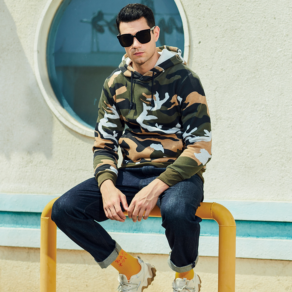 Man in camouflage hoodie en zonnebril zittend op gele reling voor rond raam. Stijlvolle streetwear, casual mode, urban look.
