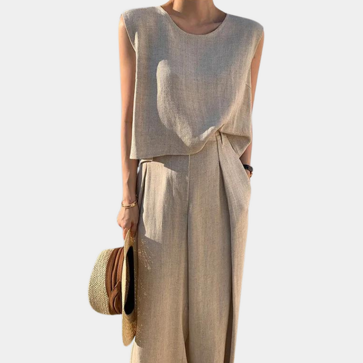 Vrouw in beige linnen zomeroutfit met mouwloze top en wijde broek, houdt een strohoed vast. Perfect voor casual en stijlvolle zomermode.