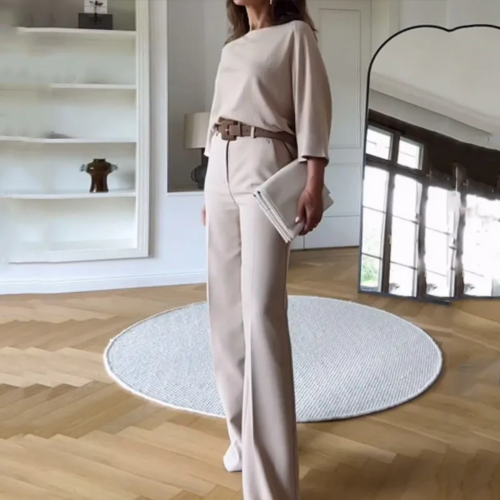 Vrouw in stijlvolle beige outfit met wijde broek en riem, staand in moderne woonkamer met houten vloer en grote spiegel. Elegante mode, minimalistisch interieur.