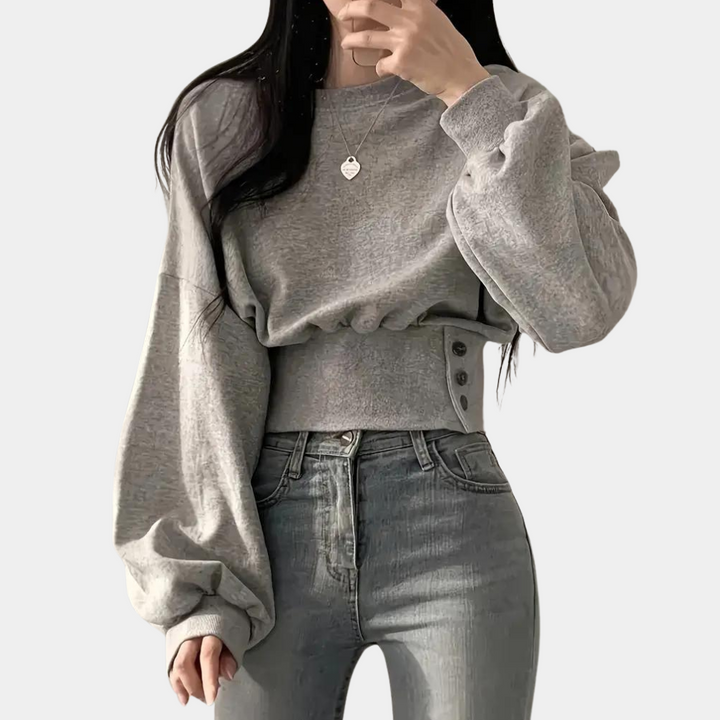 Grijze cropped trui met lange mouwen en knoopdetails, gedragen door een model. Trendy damesmode, casual stijl, comfortabele pasvorm.