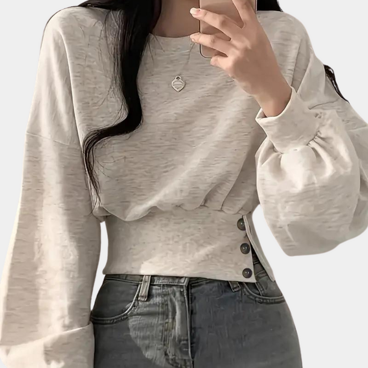 Vrouw in lichtgrijze trui met ballonmouwen en knoopdetails, draagt een ketting. Modieuze casual outfit, ideaal voor herfst- en wintermode.