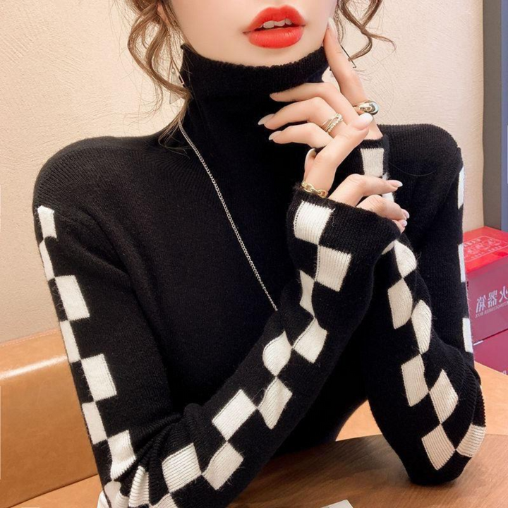 Vrouw in zwarte coltrui met zwart-wit geblokte mouwen, zittend aan tafel. Modieuze dameskleding, herfst-winter stijl, trendy design.