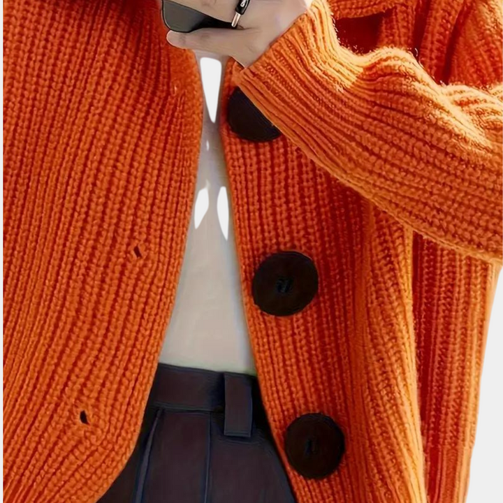 Oranje gebreid vest met grote zwarte knopen, gedragen door persoon met witte blouse en donkerblauwe broek. Modieuze herfstkleding.