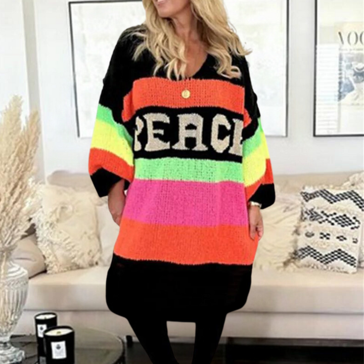 Vrouw draagt kleurrijke oversized trui met "PEACE" opdruk, zittend op beige bank in moderne woonkamer. Trendy damesmode, herfstkleding, comfortabele stijl.