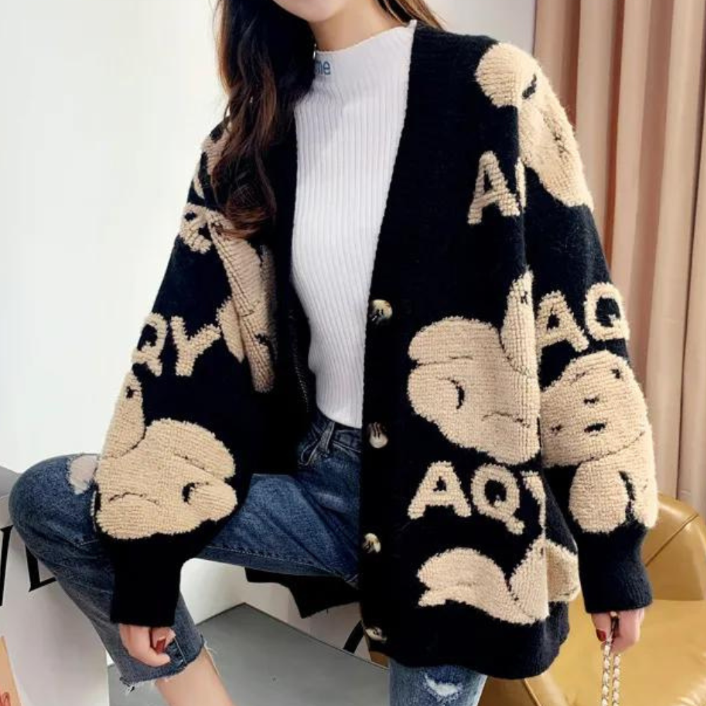 Zwarte gebreide damesvest met beige cartoonprint en knopen, gecombineerd met een witte coltrui en blauwe jeans. Trendy herfstmode.