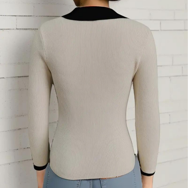 Beige geribbelde damestrui met zwarte kraag en manchetten, driekwart mouwen, getoond op een model tegen een witte bakstenen muur.