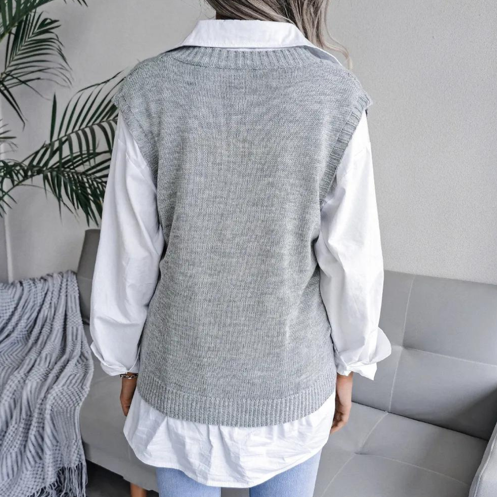 Grijze gebreide spencer over witte blouse, gedragen door vrouw, zittend op grijze bank met plant op achtergrond. Stijlvolle damesmode outfit.