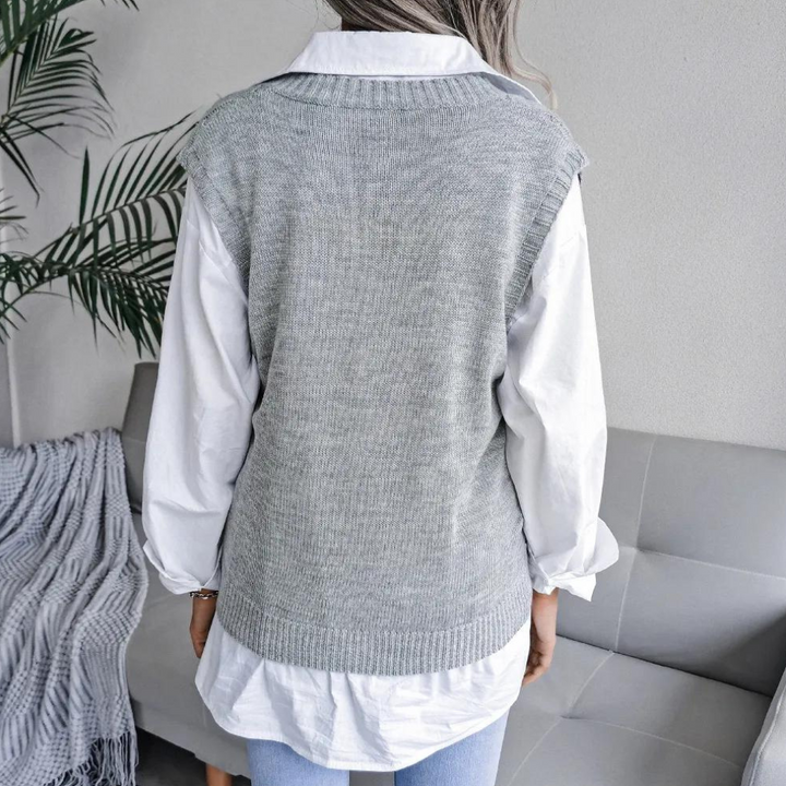 Grijze gebreide spencer over witte blouse, gedragen door vrouw, zittend op grijze bank met plant op achtergrond. Stijlvolle damesmode outfit.