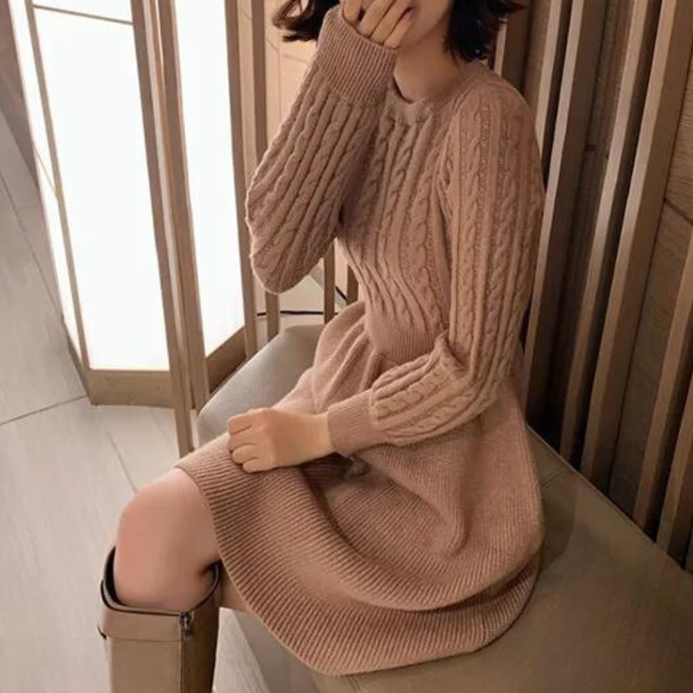 Vrouw in beige gebreide jurk met kabelpatroon, zittend op een bank. Modieuze herfstkleding, comfortabele en stijlvolle damesmode.