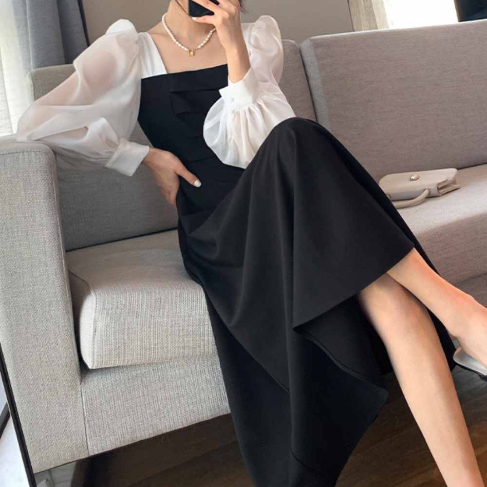 Elegante zwarte jurk met pofmouwen en witte blouse, zittend op grijze bank. Modieuze dameskleding, stijlvolle outfit voor speciale gelegenheden.