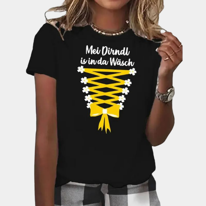 Vrouw draagt zwart T-shirt met gele dirndl-print en tekst "Mei Dirndl is in da Wäsch". Oktoberfest mode, casual kleding, grappige opdruk.