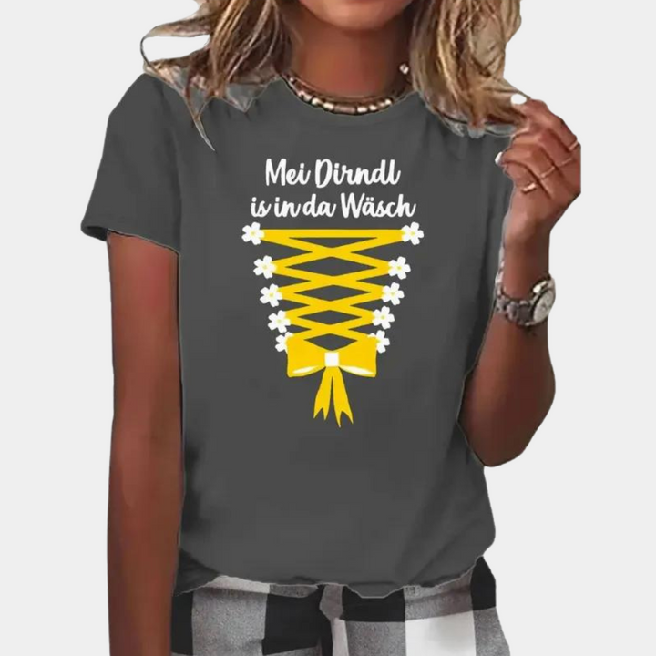 Grijs T-shirt met gele dirndl-print en tekst "Mei Dirndl is in da Wäsch", gedragen door vrouw. Perfect voor Oktoberfest, casual mode.