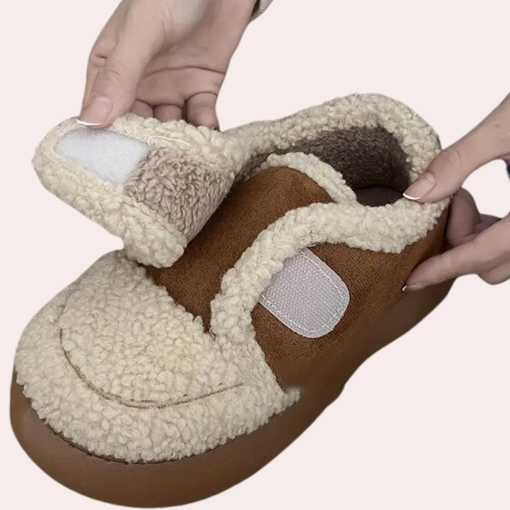 Handen houden een bruine en beige pluche pantoffel met klittenbandsluiting, ideaal voor wintercomfort en warme voeten.