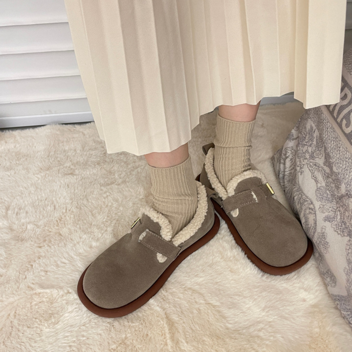 Grijze suède pantoffels met schapenvacht voering, gedragen met beige sokken op een crèmekleurig tapijt. Comfortabele wintermode voor thuis.