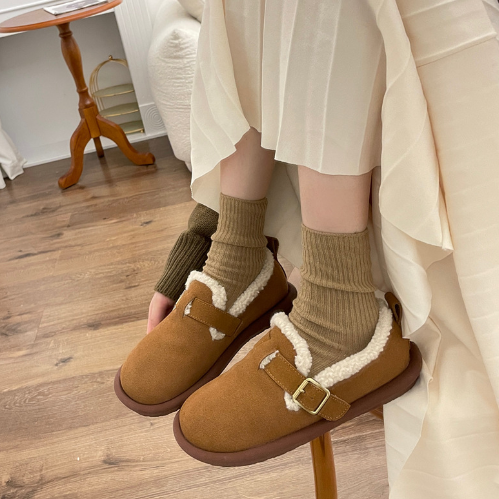 Vrouw met bruine schapenvacht pantoffels en beige sokken op houten vloer. Comfortabele wintermode, warme huispantoffels, trendy dameskleding.