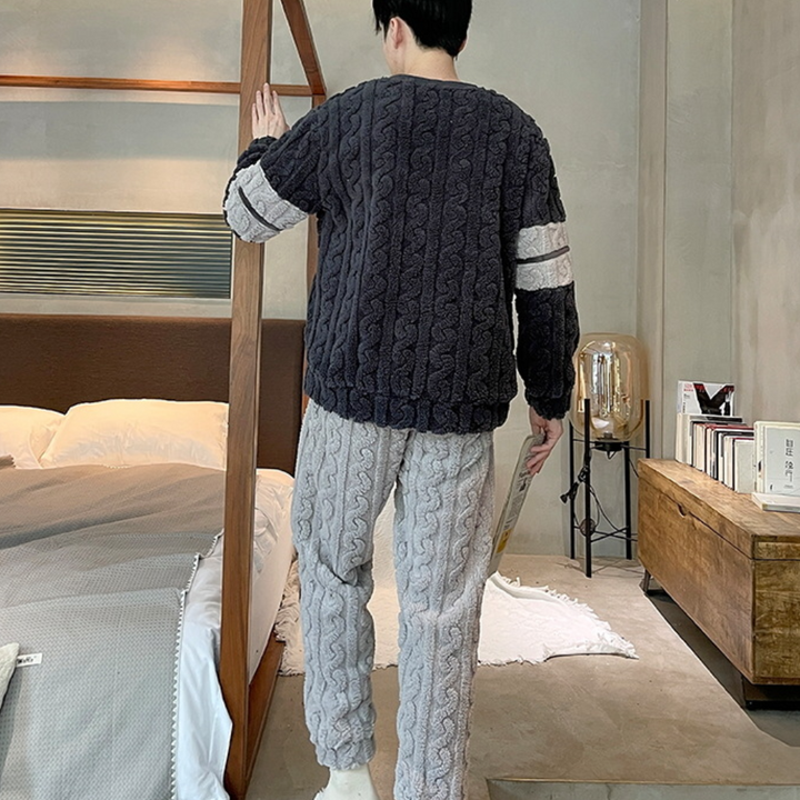 Man in grijze en zwarte kabeltrui en broek, staand in moderne slaapkamer met houten bedframe en nachtkastje. Comfortabele winterkleding, huiselijke sfeer.