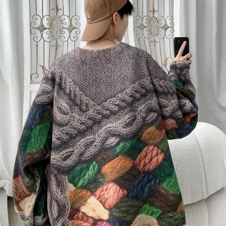 Man in oversized kleurrijke gebreide trui met kabelpatroon, bruine pet, smartphone in hand, voor decoratief hek. Trendy herenkleding, herfstmode.