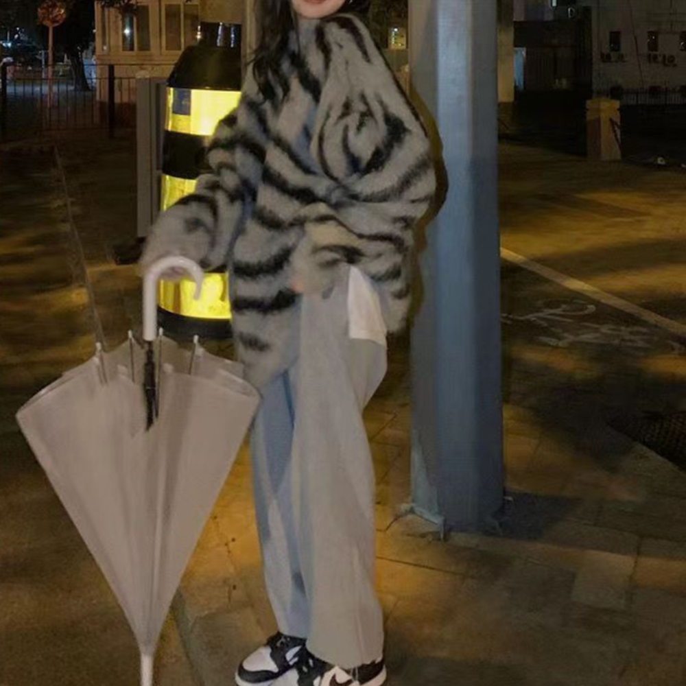 Grijze oversized bontjas met zwart-wit strepen, gedragen door persoon met grijze broek en sneakers, naast opgevouwen paraplu op straat.