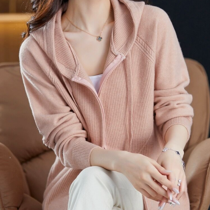 Vrouw in roze gebreid vest met capuchon, zittend op een bruine bank. Modieuze dameskleding, comfortabele casual stijl.