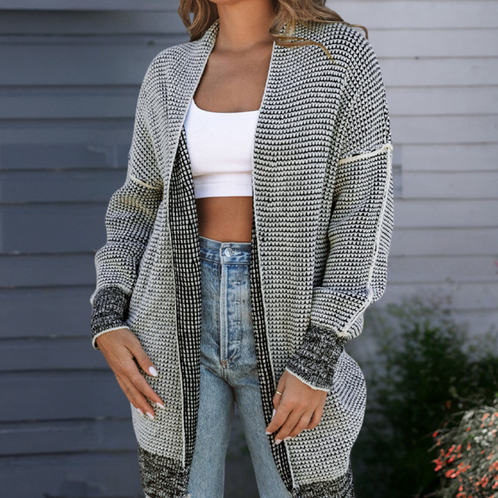 Grijze gebreide damesvest, open voorzijde, gedragen over witte crop top en blauwe jeans. Stijlvolle herfstmode, casual en comfortabel.