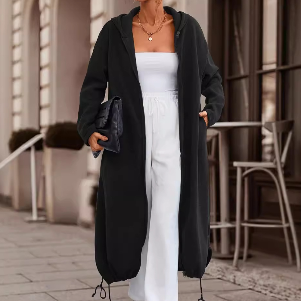 Vrouw in zwarte lange jas en witte broek, met zwarte clutch, loopt op trottoir voor café. Stijlvolle herfstmode, stedelijke achtergrond.