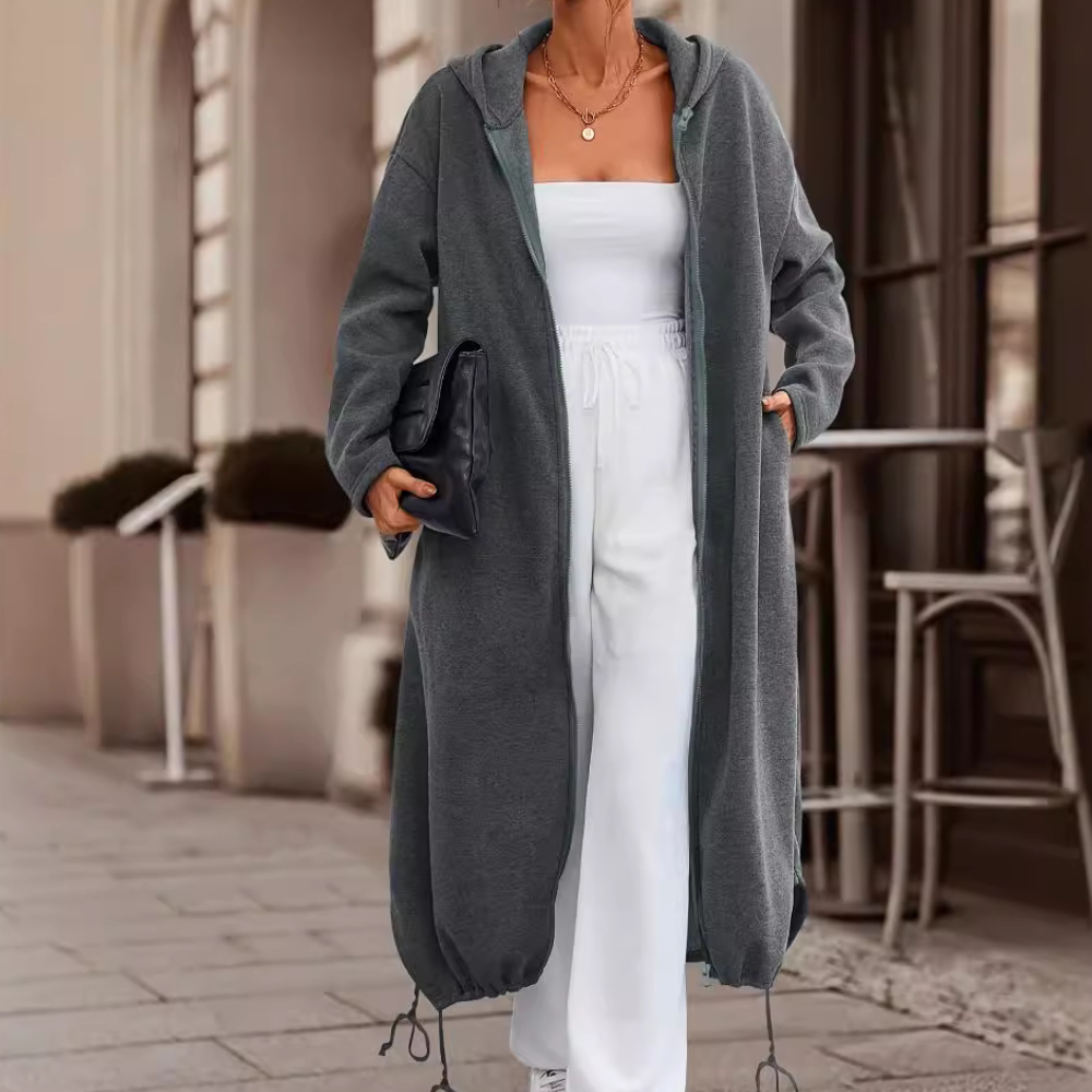 Vrouw in lange, donkergrijze hoodiejas met capuchon, witte strapless top en broek, houdt zwarte clutch vast. Stijlvolle straatmode, herfstkleding.