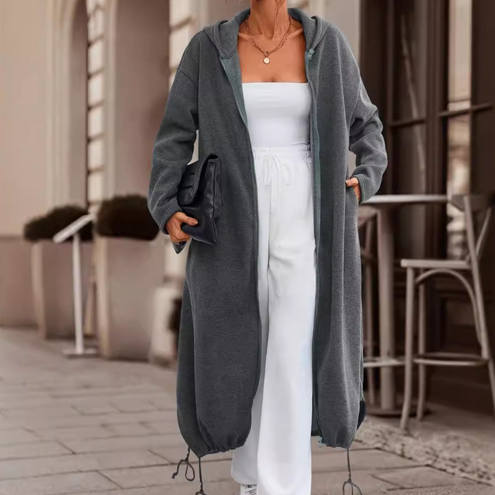 Vrouw in lange, donkergrijze hoodiejas met capuchon, witte strapless top en broek, houdt zwarte clutch vast. Stijlvolle straatmode, herfstkleding.