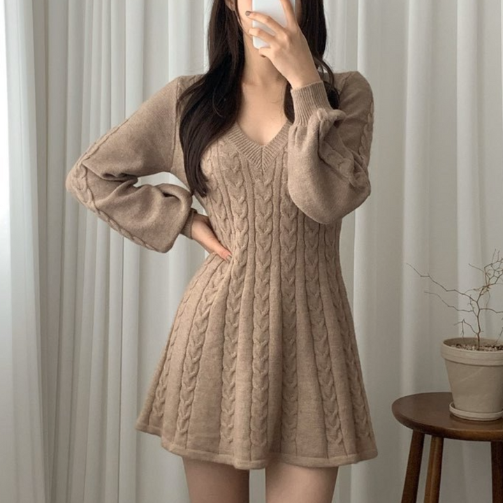 Vrouw in beige gebreide jurk met kabelpatroon, lange mouwen en V-hals, poseert voor witte gordijnen. Trendy herfstmode, comfortabele dameskleding.