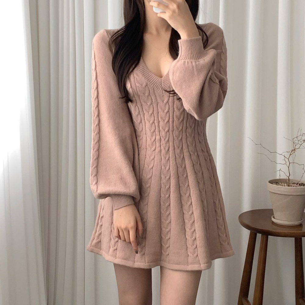 Vrouw in roze gebreide jurk met kabelpatroon, lange mouwen en V-hals, voor witte gordijnen. Trendy damesmode, herfst/winter outfit.