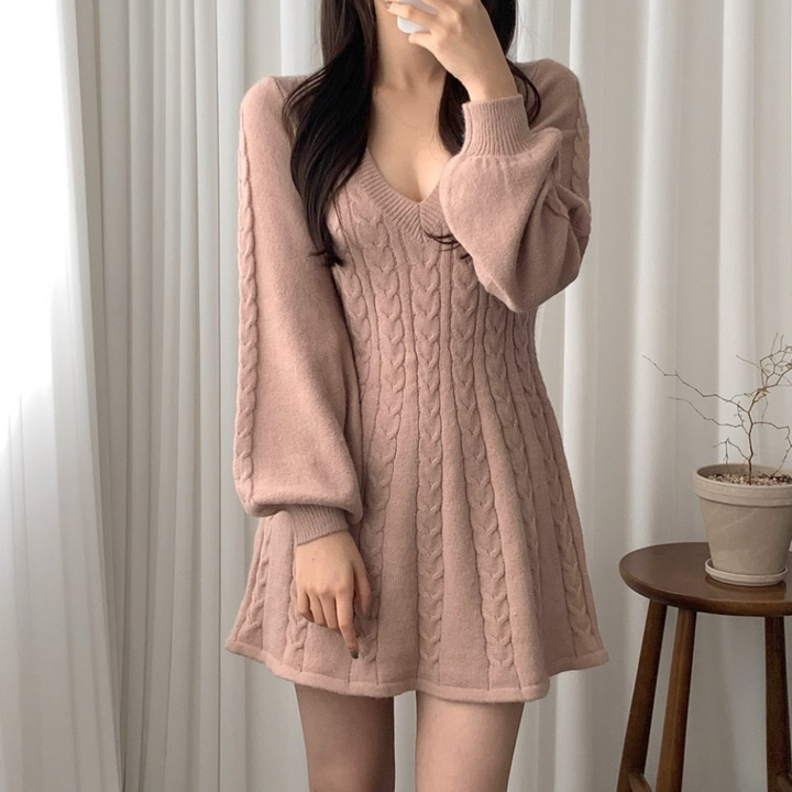 Vrouw in roze gebreide jurk met kabelpatroon, lange mouwen en V-hals, voor witte gordijnen. Trendy damesmode, herfst/winter outfit.
