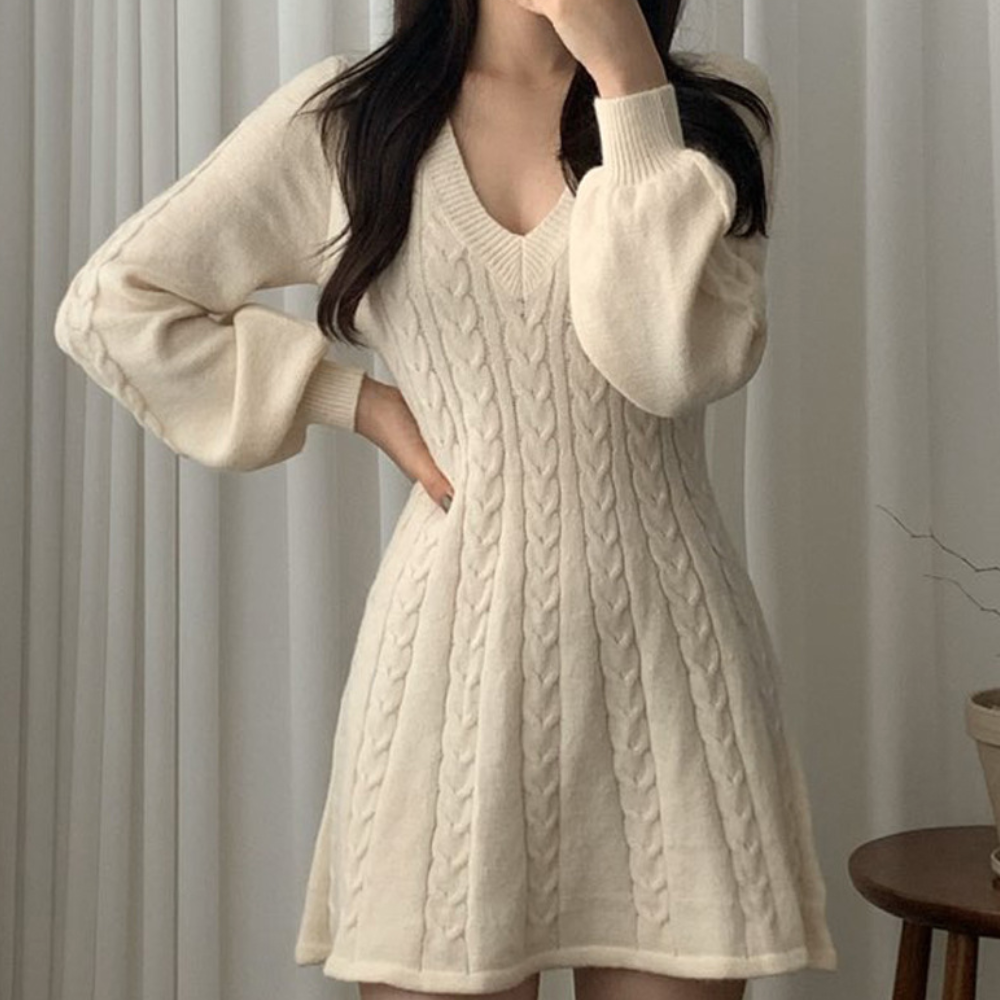 Vrouw in crèmekleurige kabeltrui jurk met V-hals, lange mouwen, voor witte gordijnen. Stijlvolle wintermode, comfortabele dameskleding.