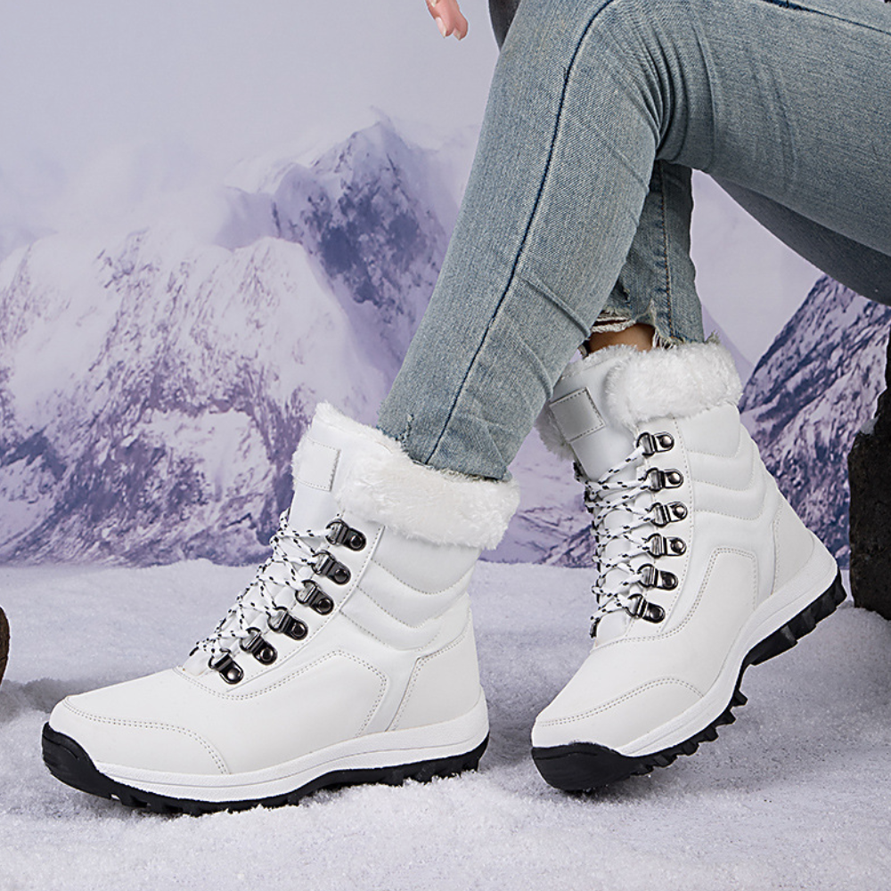 Witte winterlaarzen met bontvoering en stevige zolen, gedragen door persoon in spijkerbroek, tegen een besneeuwd berglandschap. Perfect voor koude weersomstandigheden.