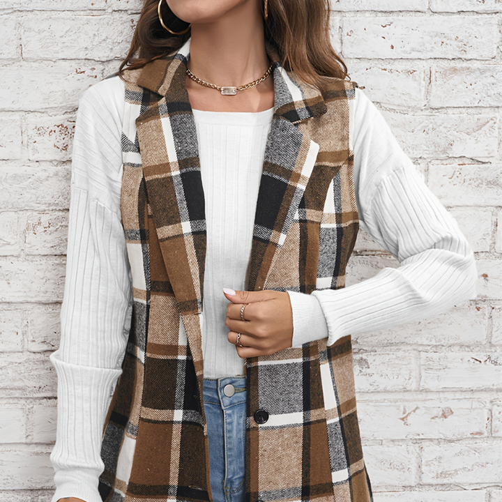 Vrouw draagt geruite bruine en zwarte lange gilet over witte trui, casual herfstmode, trendy dameskleding, stijlvolle outfit tegen bakstenen muur.