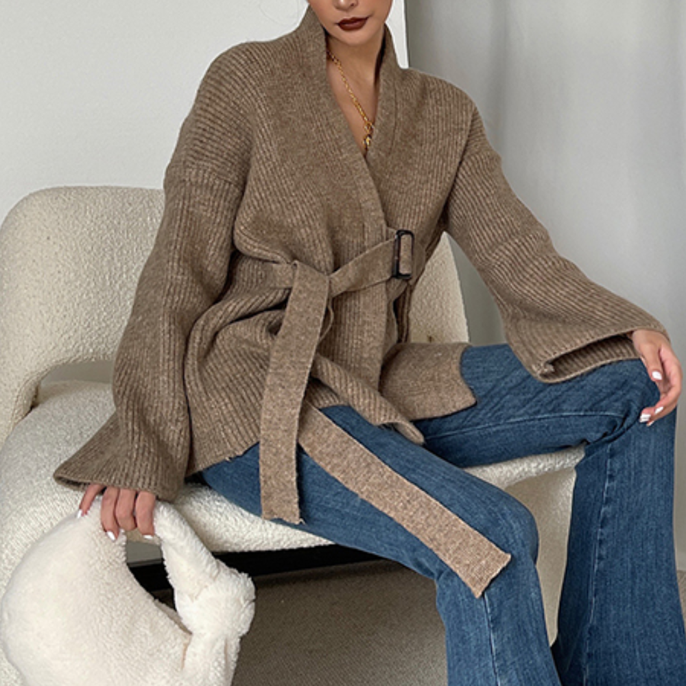 Vrouw in beige gebreid vest met riem, zittend op witte stoel, draagt blauwe jeans en houdt een witte pluchen tas vast. Modieuze herfstkleding.