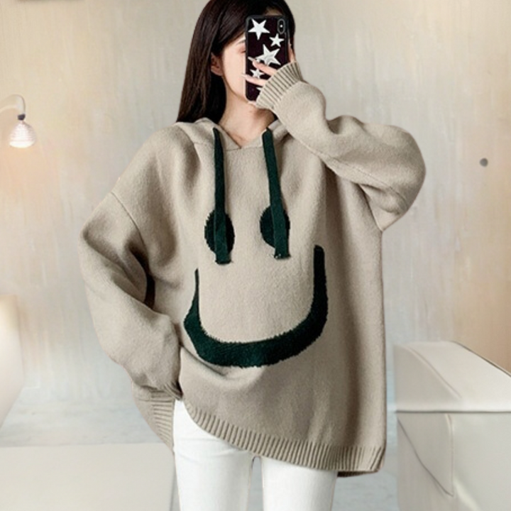 Vrouw in oversized beige trui met lachend gezicht ontwerp, capuchon en groene accenten, maakt selfie. Trendy mode, casual winterkleding.