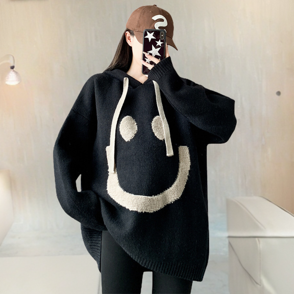 Vrouw in zwarte oversized trui met smiley-gezicht, draagt een bruine pet en houdt een telefoon vast. Trendy casual mode, comfortabele winterkleding.