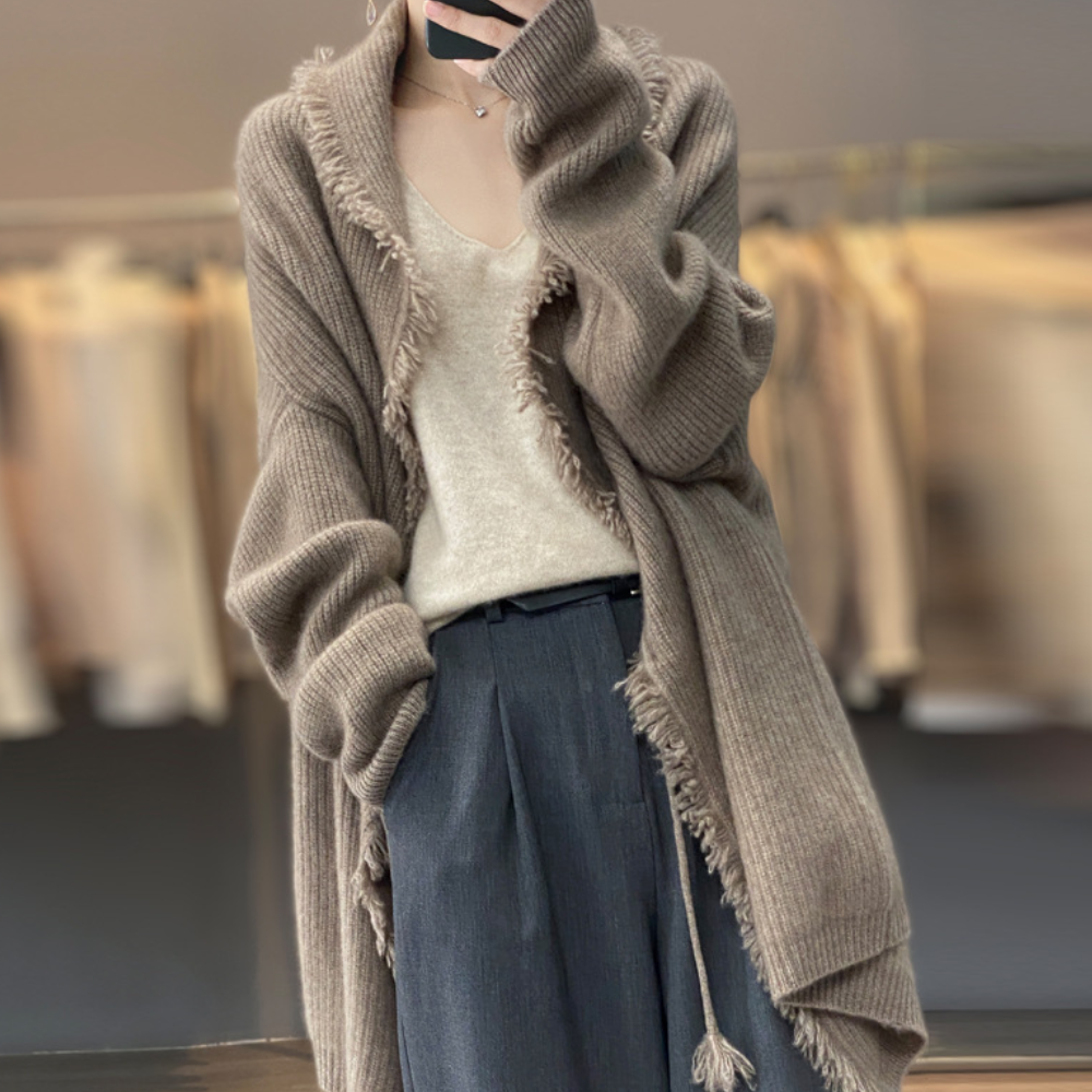 Vrouw in oversized camel-kleurige gebreide cardigan met franjes, gecombineerd met een crèmekleurige top en donkergrijze broek. Modieuze herfstoutfit.