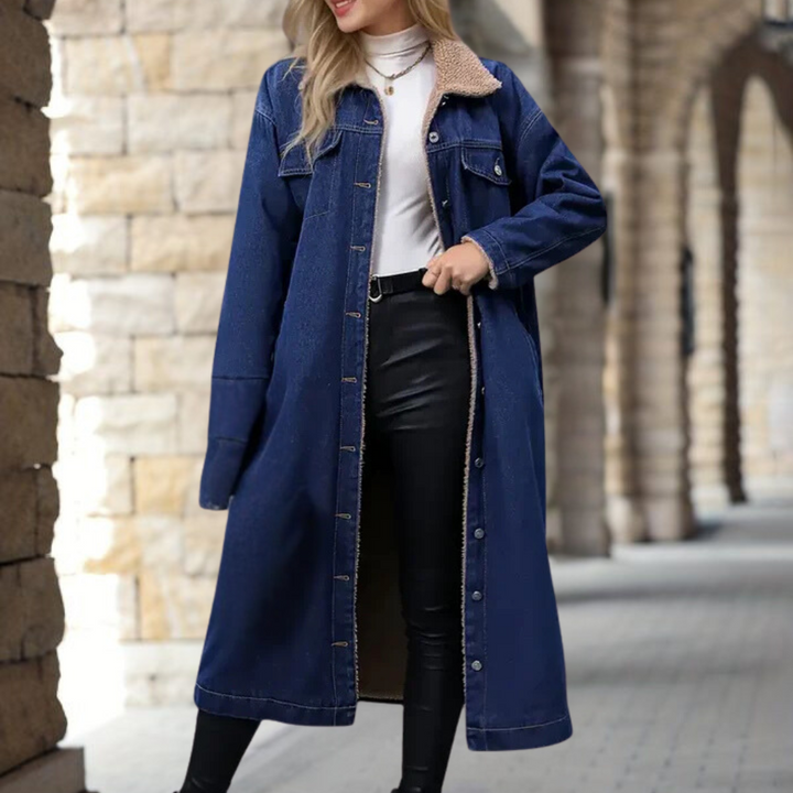 Vrouw in lange, donkerblauwe spijkerjas met knopen, witte top en zwarte leren broek. Modieuze herfstkleding, stedelijke achtergrond.