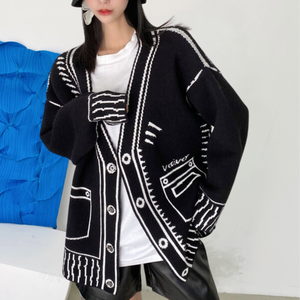Vrouw draagt zwarte oversized cardigan met witte grafische patronen en knopen, gecombineerd met witte top en zwarte leren shorts. Modieuze herfstkleding.