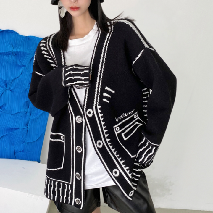 Vrouw draagt zwarte oversized cardigan met witte grafische patronen en knopen, gecombineerd met witte top en zwarte leren shorts. Modieuze herfstkleding.