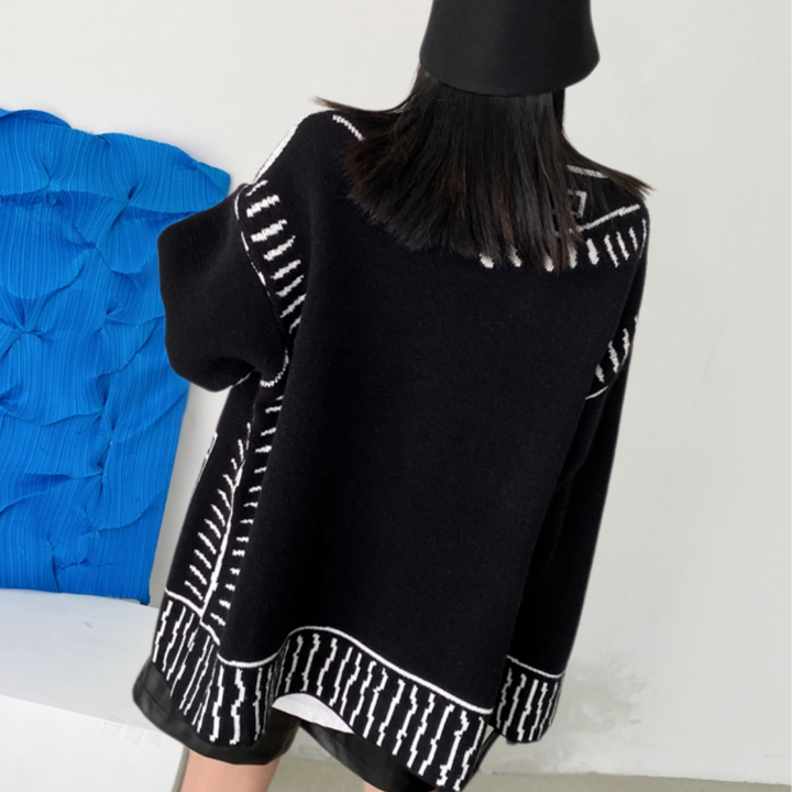 Zwarte oversized trui met witte geometrische patronen, gedragen door een persoon met lang haar en een zwarte hoed, tegen een blauwe achtergrond.