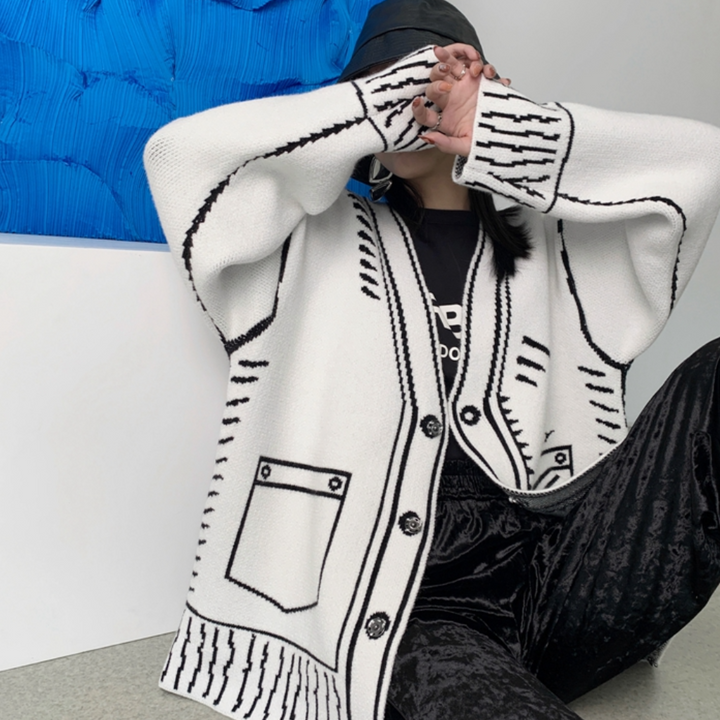 Vrouw draagt witte gebreide cardigan met zwarte strepen en zakken, gecombineerd met zwarte broek en hoed. Modieuze dameskleding, casual stijl.